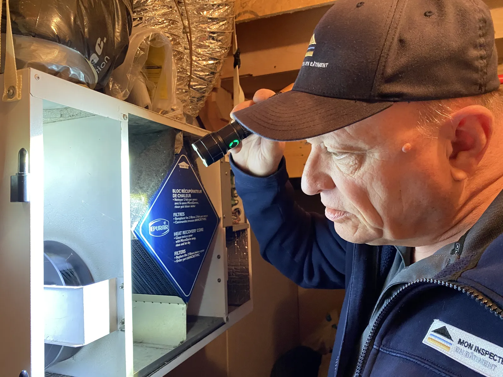 Christophe Muller inspectant un échangeur d'air à la lampe frontale lors d'une inspection de bâtiment à Auteuil, Laval.